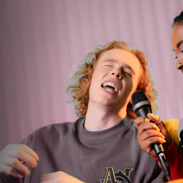 Young man with curly red hair singing a high note with his eyes closed, a friend holding the mic for him