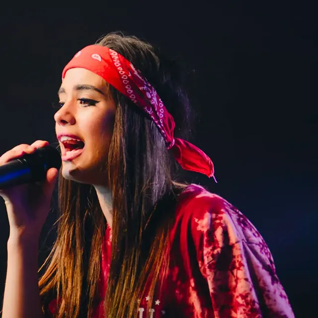 Young woman in a red bandana singing into a handheld microphone on a dark stage