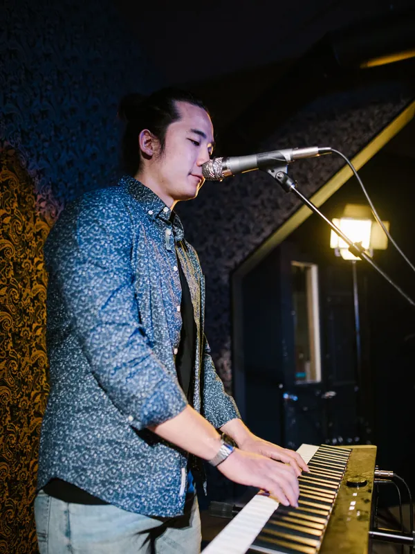 Young man singing into a microphone while playing a keyboard in a dim room