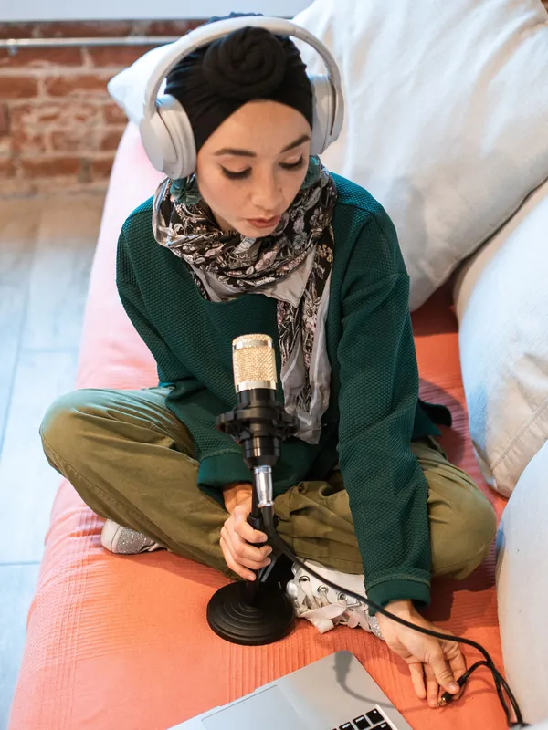 Woman in a headscarf and headphones recording at home with a microphone and laptop