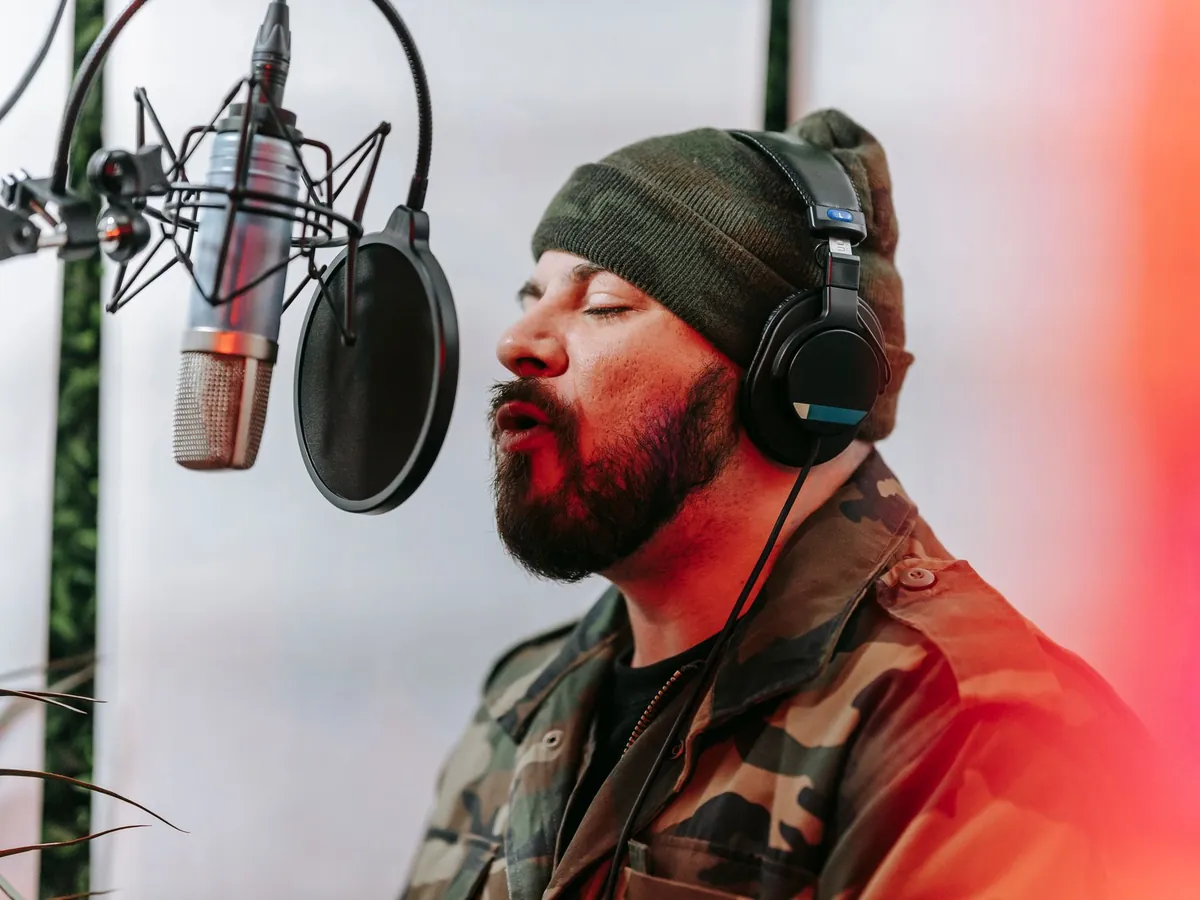Bearded man in a beanie and headphones holding a long note with his eyes closed at a studio microphone